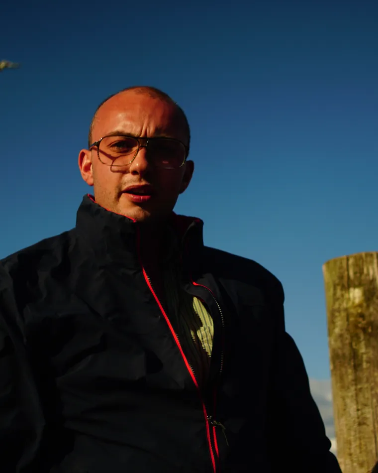 Felix Maynard outdoors on the Norfolk coast, mid-sentence, a weathered mooring post behind him against a clear evening sky.
